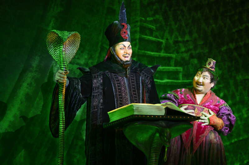 Anand Nagraj as Jafar and Aaron Choi as Iago in the North American Tour of Aladdin. Jafar, dressed in a long black robe, holds a golden snake staff as he stands over a book on a stand. Iago, dressed in a red robe, stands next to Jafar looking at him.