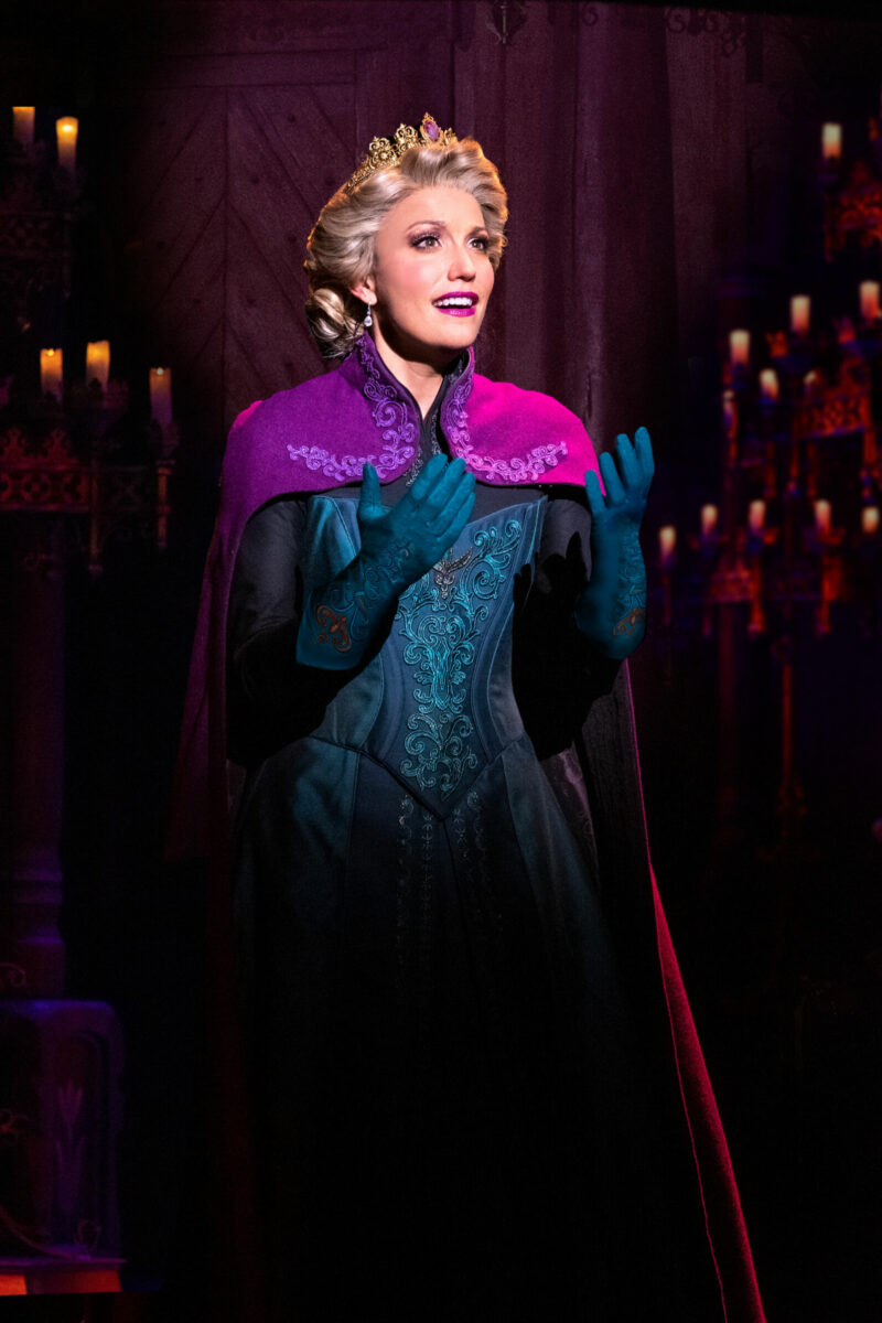 Caroline Bowman as Elsa in Frozen North American Tour. Queen Elsa, wearing a blue corset and skirt with matching gloves and a purple shawl, stands palms facing up as she looks into the distance. Many dimly lit candles are behind her.