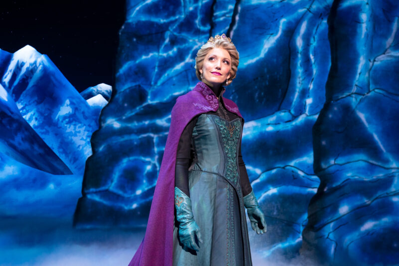 Caroline Bowman as Elsa in Frozen North American Tour. Queen Elsa, wearing a blue corset and skirt with matching gloves and a purple shawl, looks off into the distance smiling.