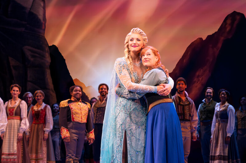 Caroline Bowman as Elsa and Lauren Nicole Chapman as Anna and Company in Frozen North American Tour. Anna and Elsa embrace and look hopefully into the distance as spectators look on.