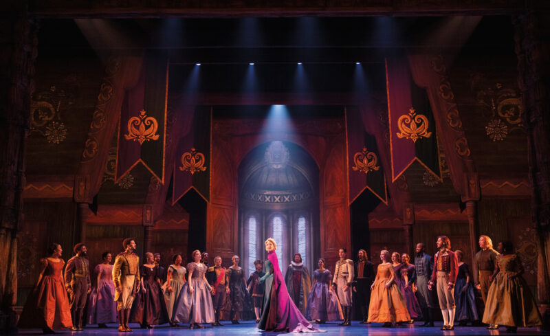 Caroline Bowman as Elsa and the Company of Frozen North American Tour. Elsa stands alone in the center of a ball as the guests surround and stare at her.