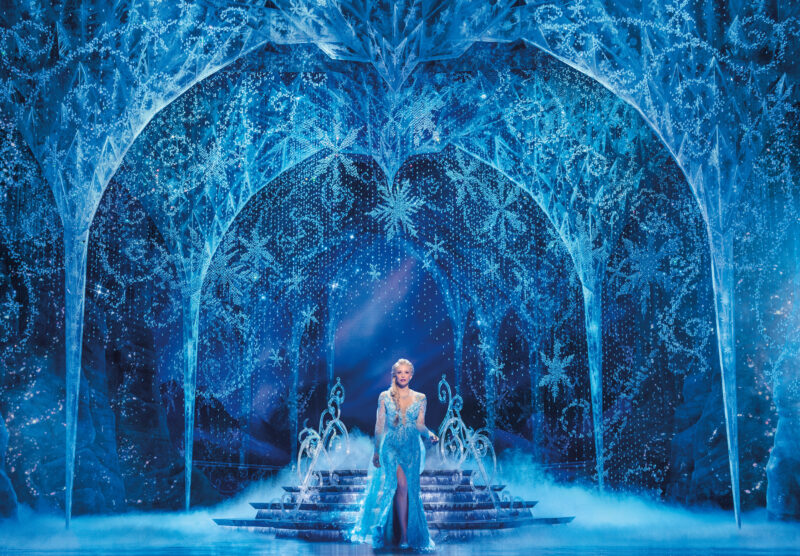 Caroline Bowman as Elsa in Frozen North American Tour. Elsa, in her signature long blue flowy dress, stands in front of a descending staircase. She is in her ice castle, with tall ceilings covered in snowflakes.