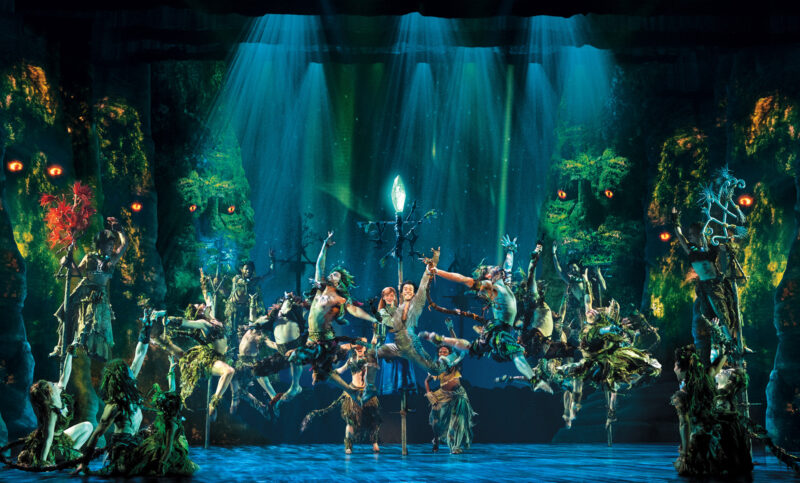 Company of the Frozen North American Tour. Dancers, adorned in grassy garments, leap into the air gracefully. Large grassy faces with glowing orange eyes watch over the dancers.