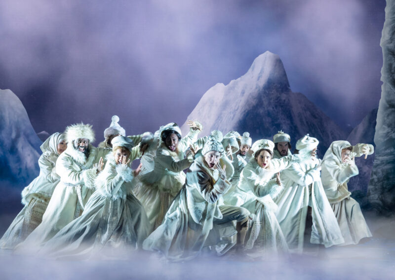 Company of the Frozen North American Tour. Several people, dressed head to toe in white, stand bunched together and leaning to one side. Some have their hands up protecting their faces. There are snowy mountains in the background.