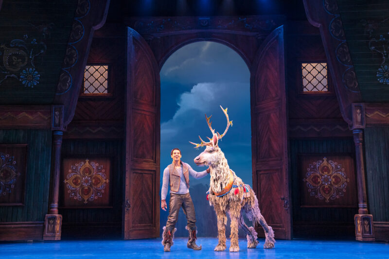 Dominic Dorset as Kristoff and Collin Baja as Sven in Frozen North American Tour. Kristoff stands next to Sven, a reindeer with antlers that make him taller than Kristoff, in front of large wooden barn doors.