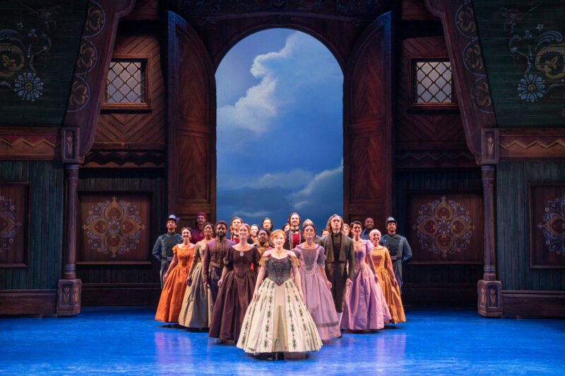 Lauren Nicole Chapman as Anna and Company in Frozen North American Tour. Anna and her guests are dressed in ball gowns and formal clothing as they stand in a triangular formation with Anna at the point.